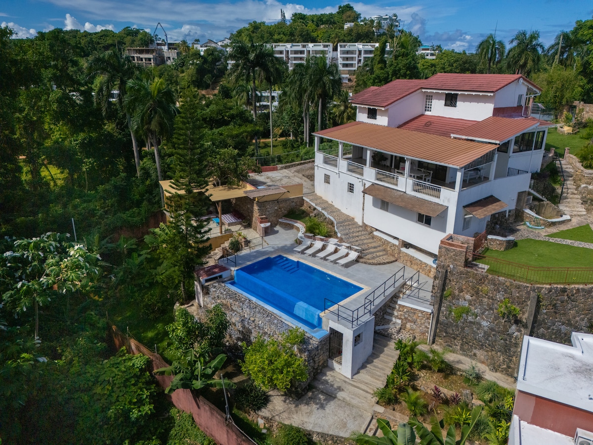 An aerial view captures a spacious property featuring a large pool surrounded by lounge chairs. Lush greenery enhances the outdoor area, and a modern structure is visible in the background, displaying a combination of stone and white walls, with a terracotta roof and balconies.