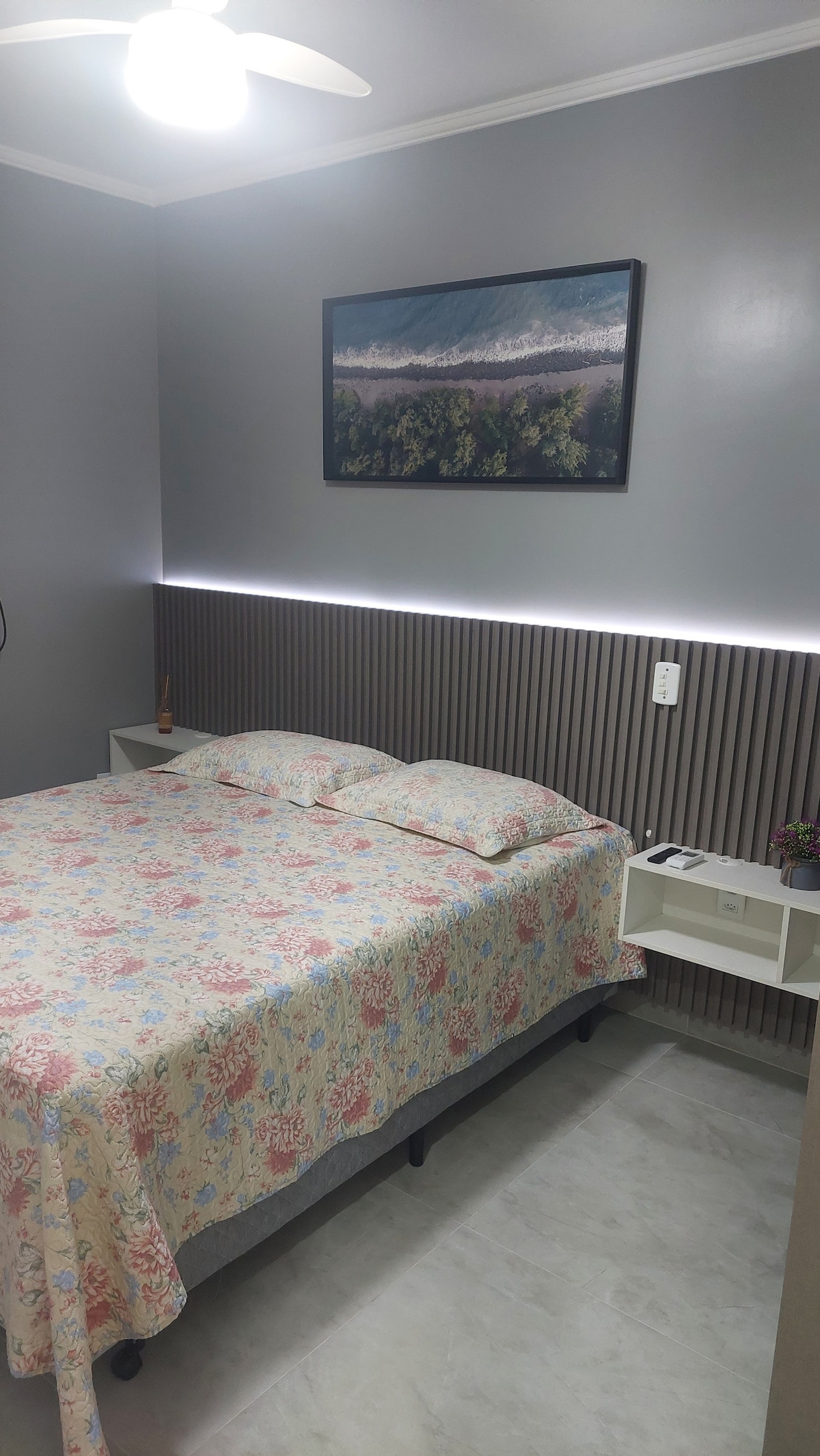 A modern bedroom showcases a spacious bed dressed in a floral patterned blanket. Two side tables are positioned on either side, complemented by soft wall lighting. A nature-themed artwork hangs above the bed, while a ceiling fan provides additional comfort.