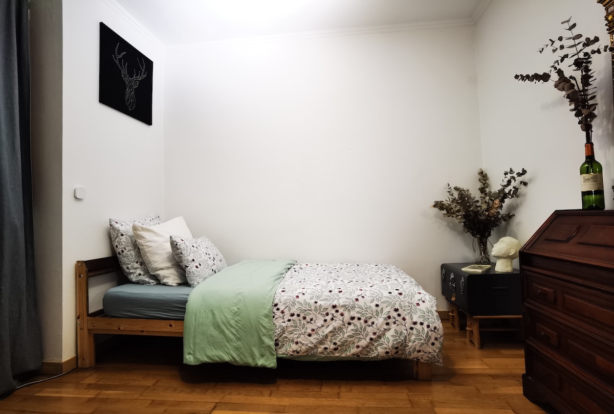 A comfortable sleeping area is featured, showcasing a wooden bed with a floral patterned duvet. Soft pillows rest against the wall, while a small nightstand holds decorative items. A wine bottle and a sculpture are placed nearby, complemented by a wall art piece depicting a deer.