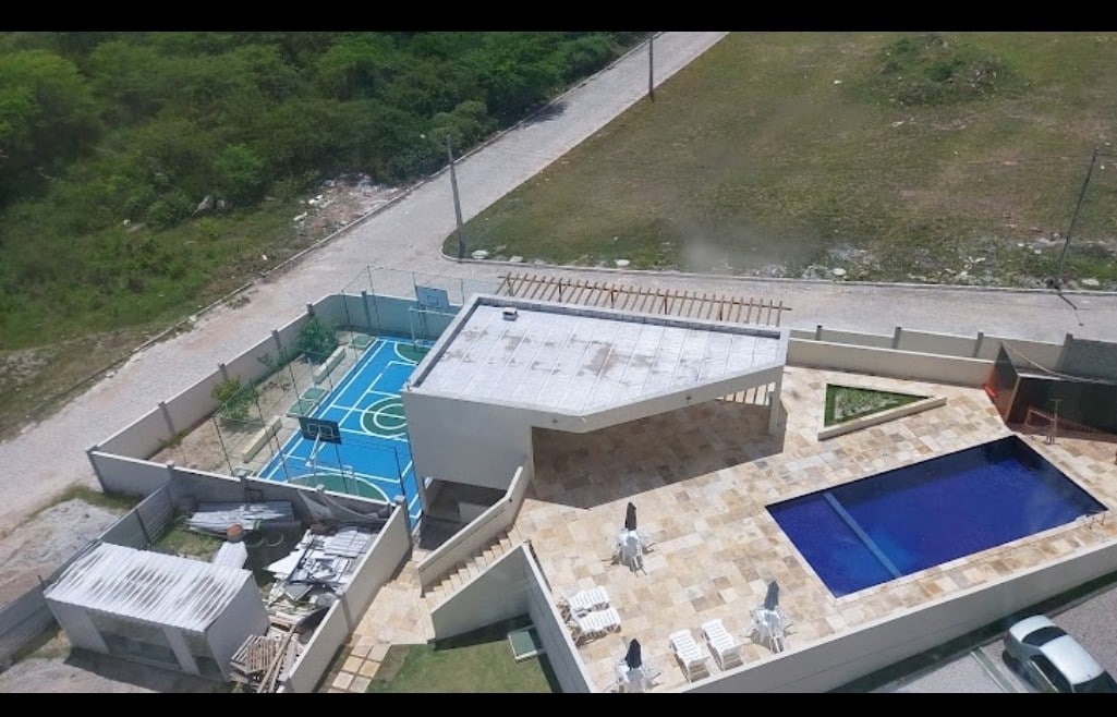 An aerial view showcases a modern property featuring a spacious swimming pool surrounded by stone tiles. Nearby, a basketball court is visible, along with an outdoor seating area. Lush greenery and an open landscape are seen in the background.