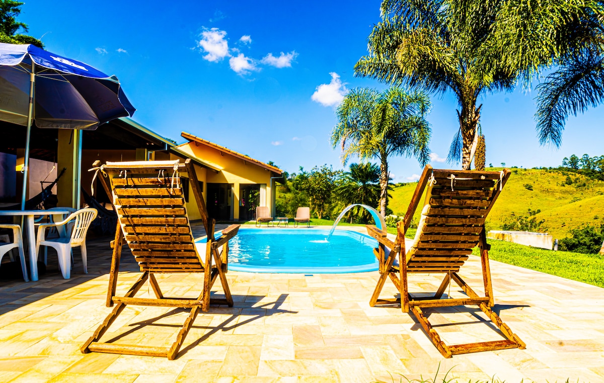 Two wooden lounge chairs are positioned by a sparkling pool surrounded by landscaped greenery. A patio table and white plastic chairs are nearby, under an umbrella. Lush hills and a clear blue sky can be seen in the background, creating a serene outdoor setting.