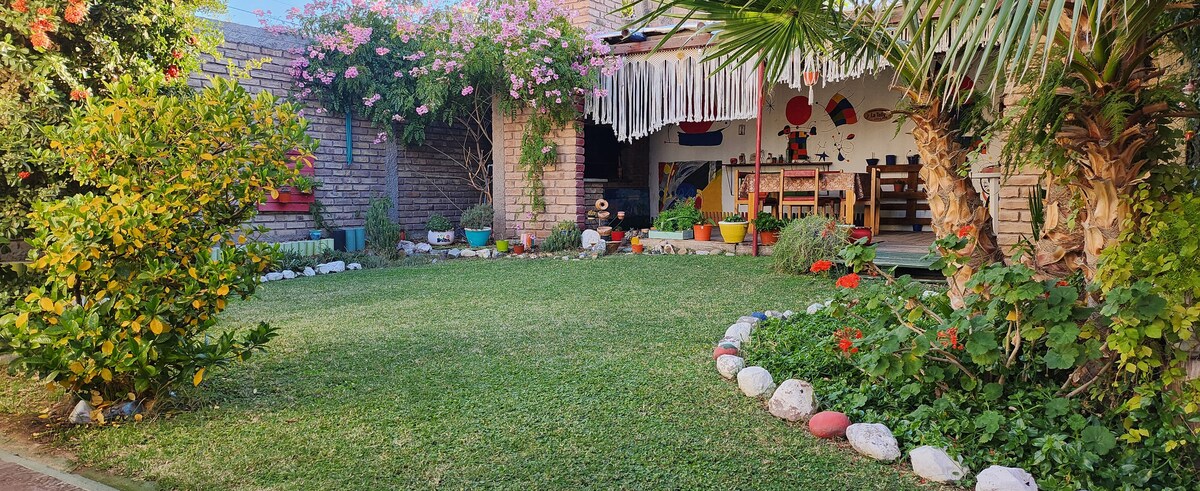 A green garden area displays neatly arranged flower beds with colorful plants and decorative stones lining the edges. A cozy patio with seating is visible in the background, surrounded by lush greenery and blooming flowers, creating a serene outdoor space.