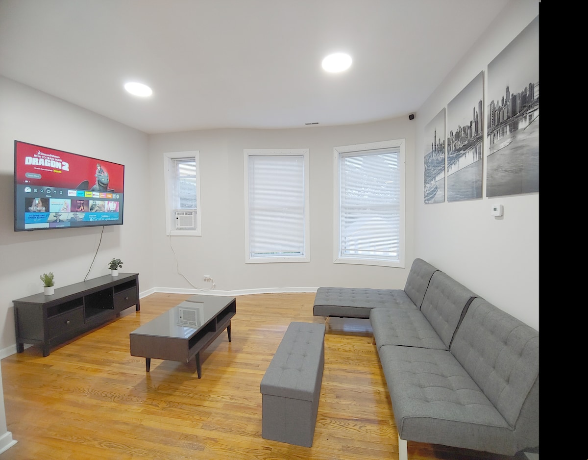 The living room features a modern sectional sofa and a sleek coffee table placed on hardwood flooring. A large wall-mounted smart TV displays various streaming options, while light enters through multiple windows, enhancing the bright and spacious atmosphere.