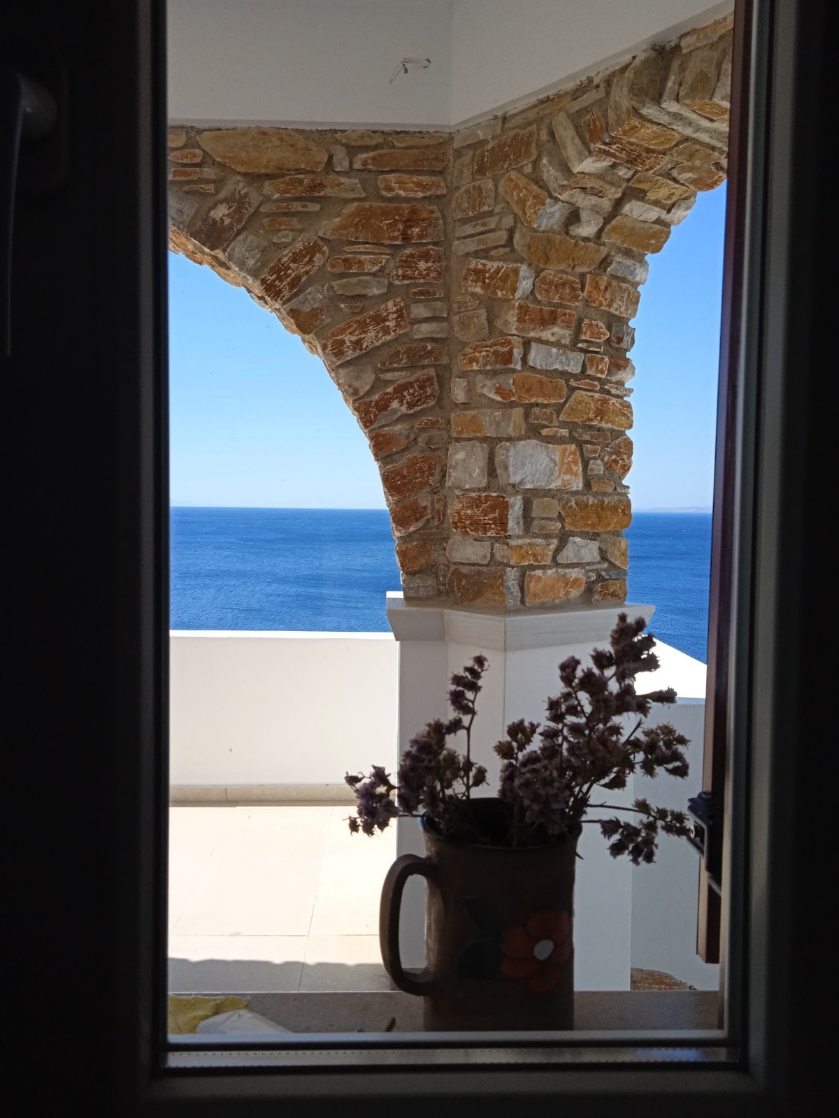A view is framed by an archway made of stone, showcasing the glistening blue sea beyond. A vase with dried flowers sits on a ledge, adding a subtle touch of decor to the scene.
