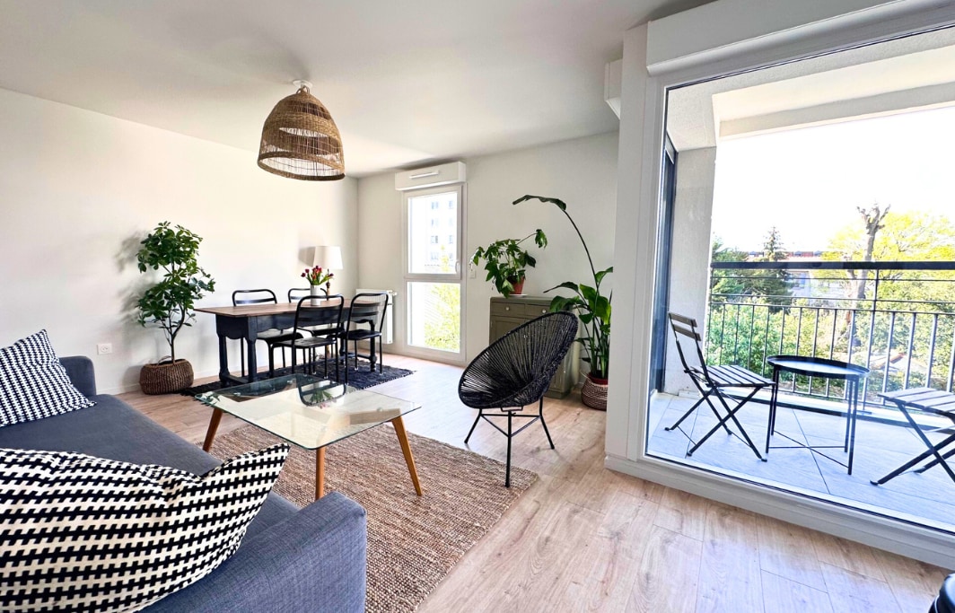 A modern living area is displayed, featuring a gray sofa and a glass coffee table on a woven rug. A dining area with a dark table and chairs is visible. Indoor plants add greenery, and large sliding doors lead to a balcony with outdoor seating.