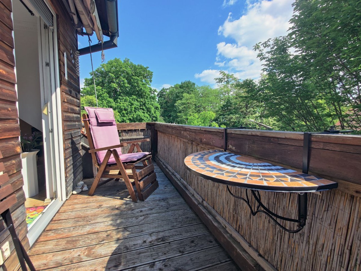 A private balcony features a hanging chair and a small foldable table with a colorful tabletop. Wooden decking surrounds the outdoor space, with greenery visible in the background and clear blue skies above.