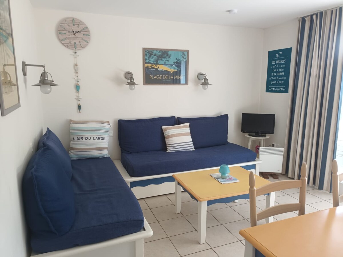A relaxing seating area showcases two blue sofas accompanied by striped cushions, arranged around a light wooden coffee table. A small television is positioned near the wall. The room is illuminated by two wall sconces, enhancing the inviting ambiance.