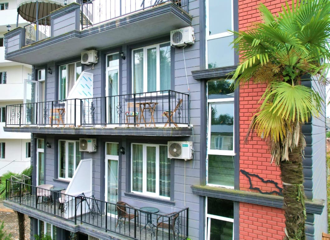 A modern three-story building is showcased, with multiple balconies featuring patio furniture. Large windows allow natural light into the rooms, and air conditioning units are visible on the exterior. A palm tree is situated nearby, adding to the tropical ambiance.
