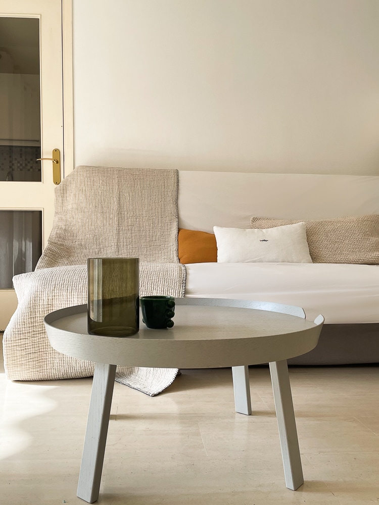 A cozy sitting area features a light-colored, modern coffee table in the foreground. A beige throw blanket drapes over a sofa, accompanied by decorative cushions in neutral tones. Soft natural light filters through a nearby glass door, highlighting the inviting space.