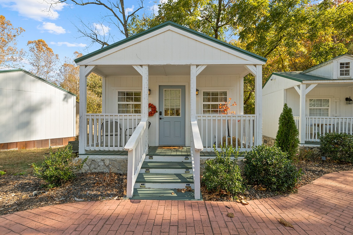 A welcoming cottage exterior features a light-colored facade with a gabled roof. A front porch is accessible by steps and adorned with white railing. The surrounding landscape includes well-maintained shrubs and trees, providing a serene natural setting.