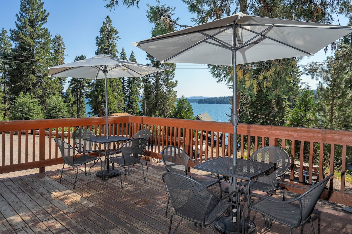 An outdoor deck features several tables with black mesh chairs, each shaded by large umbrellas. The tranquil lake can be seen in the background, surrounded by tall trees, creating a serene outdoor dining environment.
