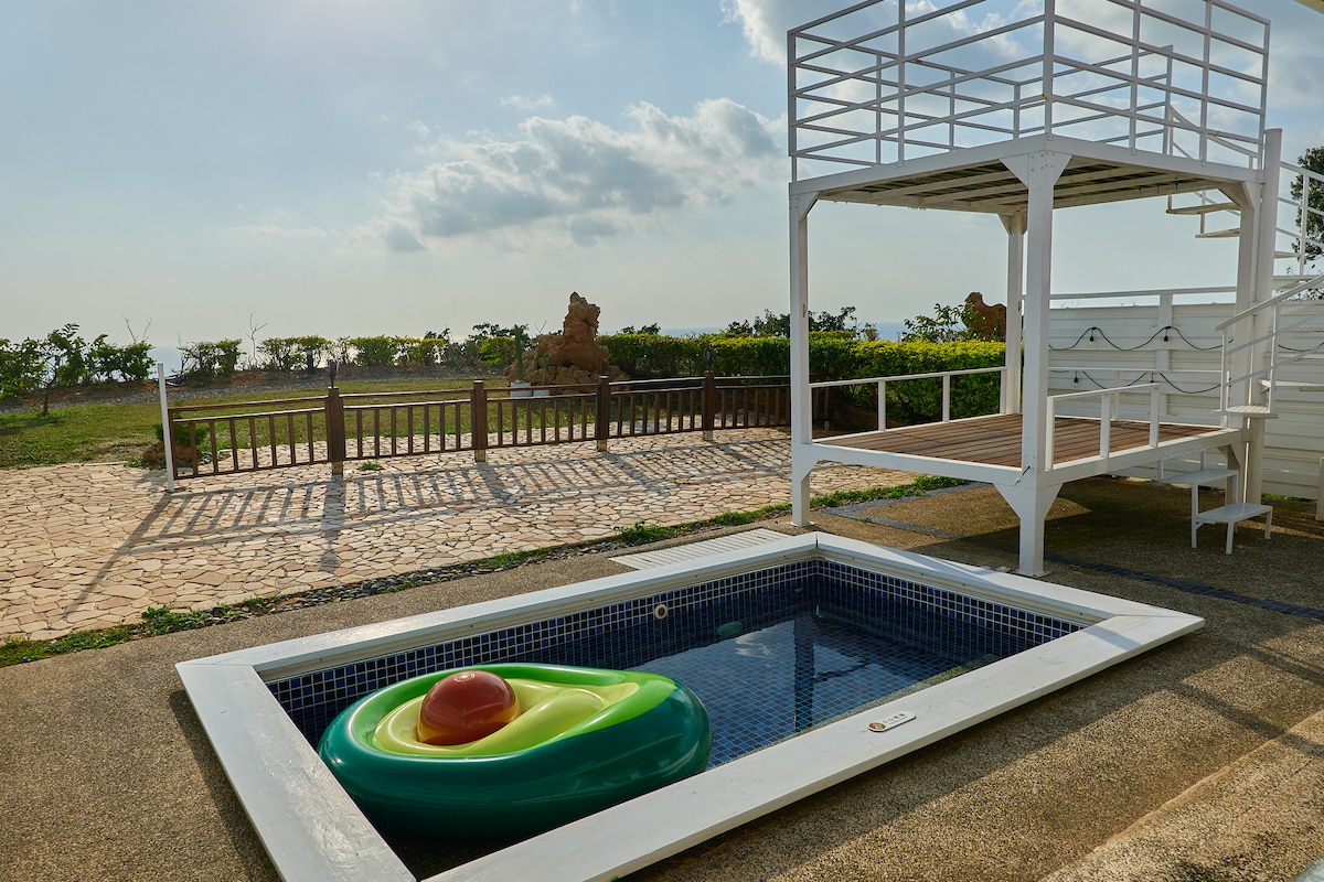 A private outdoor area features a small pool with a colorful inflatable float resting on the water's surface. Nearby, a two-level lounging structure overlooks a scenic landscape, with grassy fields and a stone-paved pathway visible in the background. Soft sunlight bathes the space.