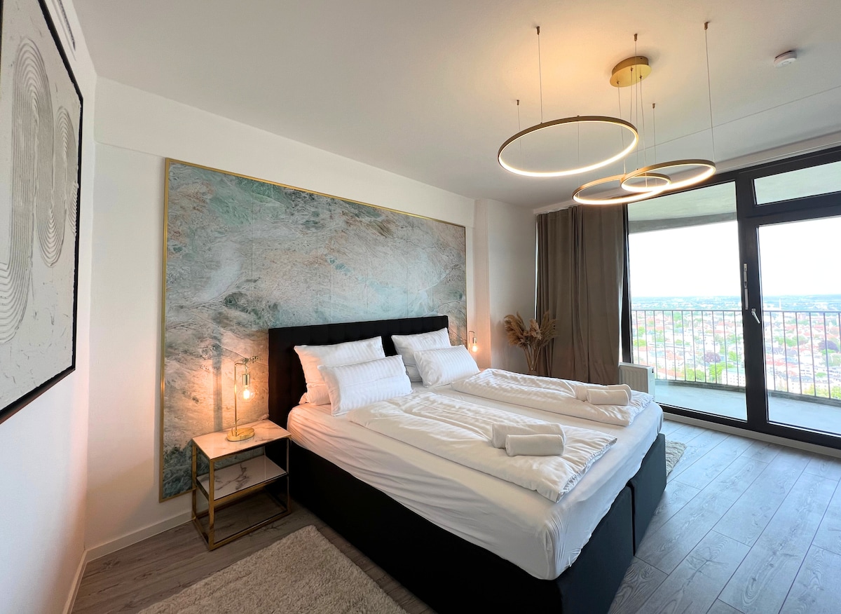 A spacious bedroom features a king-size bed adorned with crisp white linens. A large wall art piece provides a modern touch above the bed. Two elegant bedside lamps illuminate the space softly, while floor-to-ceiling windows showcase views of the cityscape.
