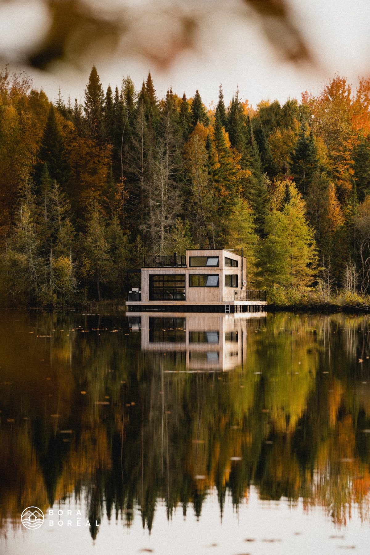 The floating chalet is surrounded by vibrant autumn foliage, with reflections of trees visible in the calm water. The modern structure features large windows that provide a striking contrast to the natural setting, and a wooden dock extends into the lake.