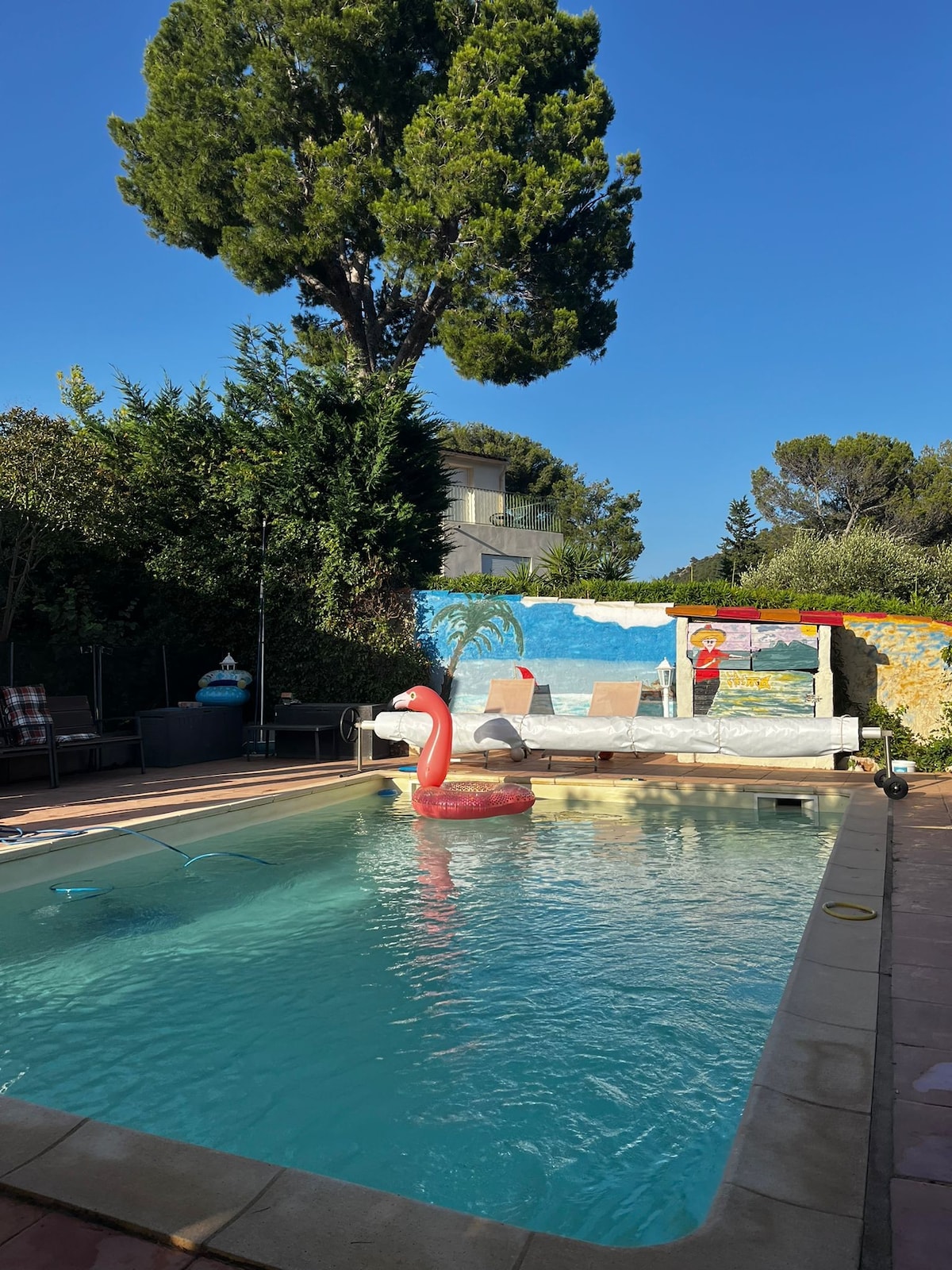 The outdoor space features a swimming pool surrounded by a paved area and lush greenery. A vibrant mural depicting a beach scene adorns the wall behind the pool. A large flamingo float adds a playful touch to the clear water. Comfortable seating is positioned nearby for relaxation.