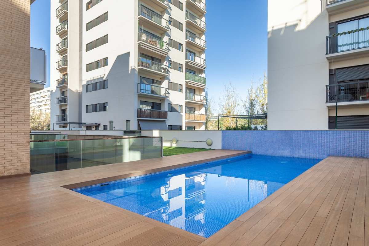A community pool is surrounded by a wooden deck and features blue water reflecting the building's exterior. Direct sunlight illuminates the area, while glass panels provide a sense of openness. Nearby greenery adds a natural touch to the urban backdrop.