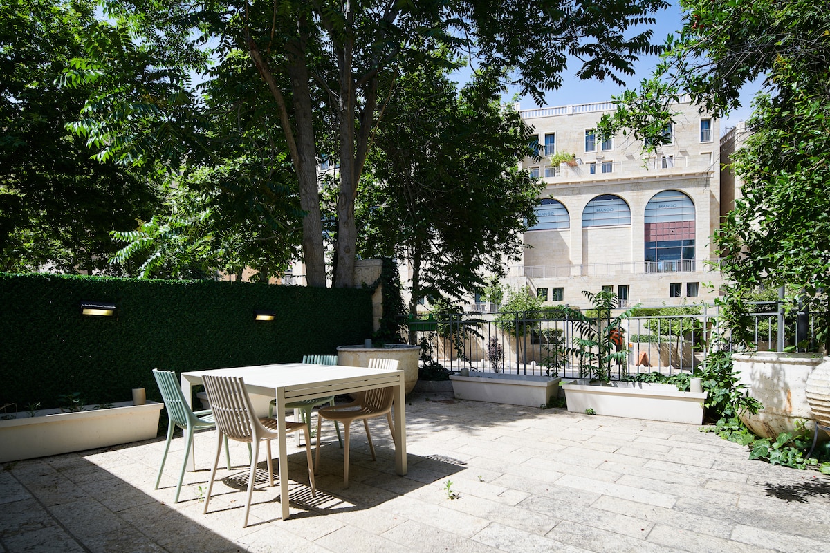 An outdoor terrace is presented with a dining table and chairs, surrounded by greenery and trees. A stone pathway leads to the table, which is set against a backdrop of a building with arched windows. The space is illuminated by natural light.