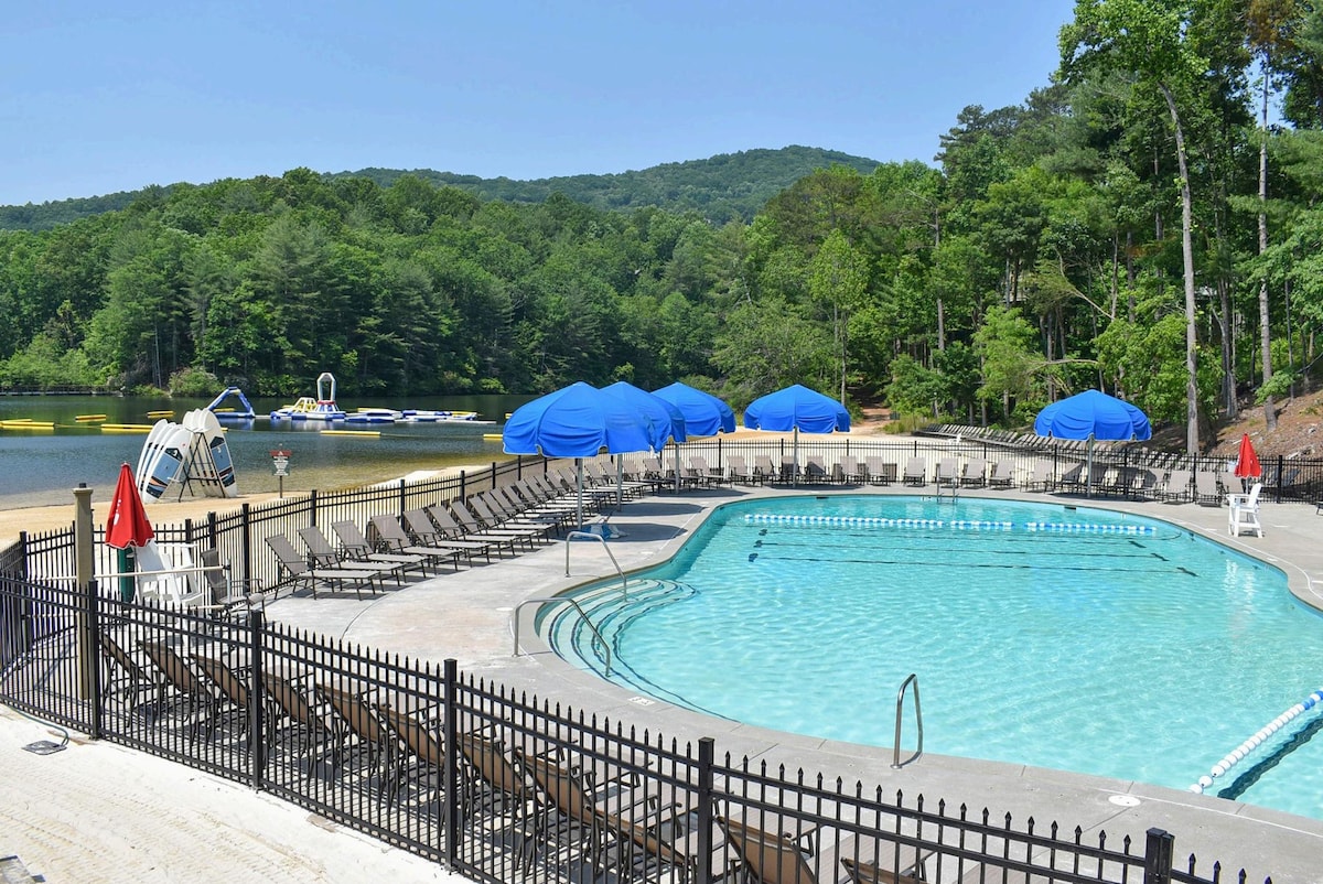 A clear blue swimming pool is surrounded by lounge chairs arranged for relaxation. Sun umbrellas provide shade for guests, while the pool's edge meets a sandy beach area. Lush green mountains rise in the background, contributing to the scenic outdoor environment.