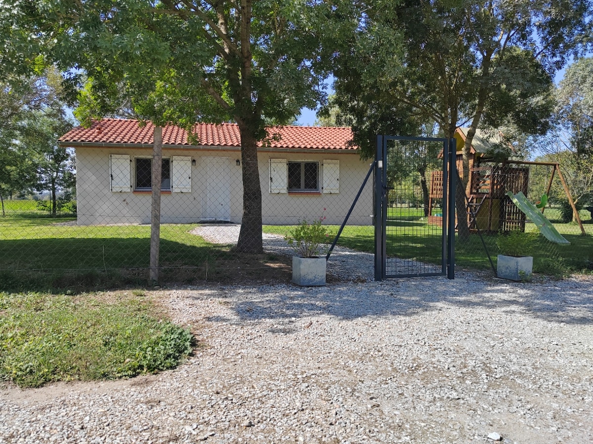 A single-story house with a terracotta roof is nestled behind a fenced yard, featuring lush greenery and a gravel path leading to the entrance. Nearby, a play structure is visible, adding to the family-friendly atmosphere of the property.