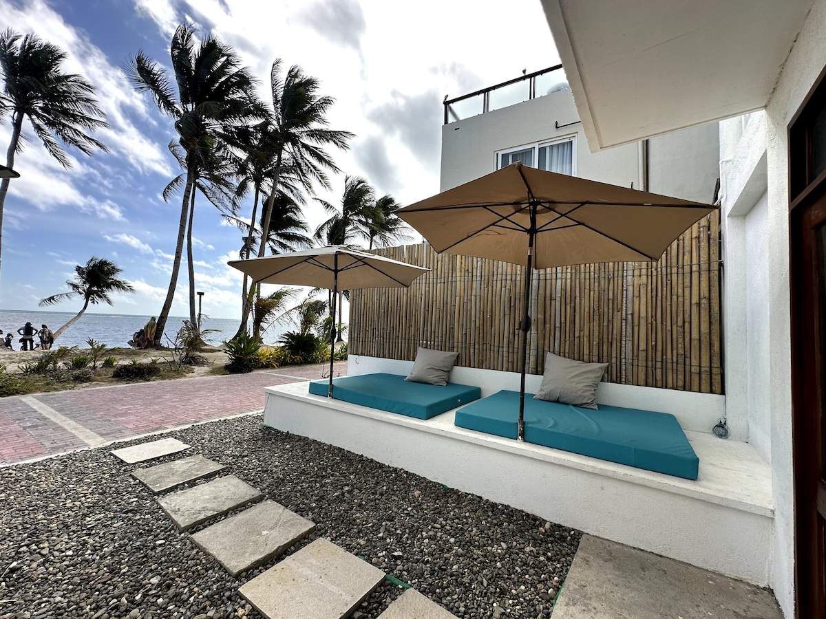 Outdoor loungers with teal cushions are positioned under large umbrellas, providing shade. The area features a landscaped pebbled ground, leading to a pathway. In the background, palm trees sway gently and the ocean is visible, enhancing the coastal setting.