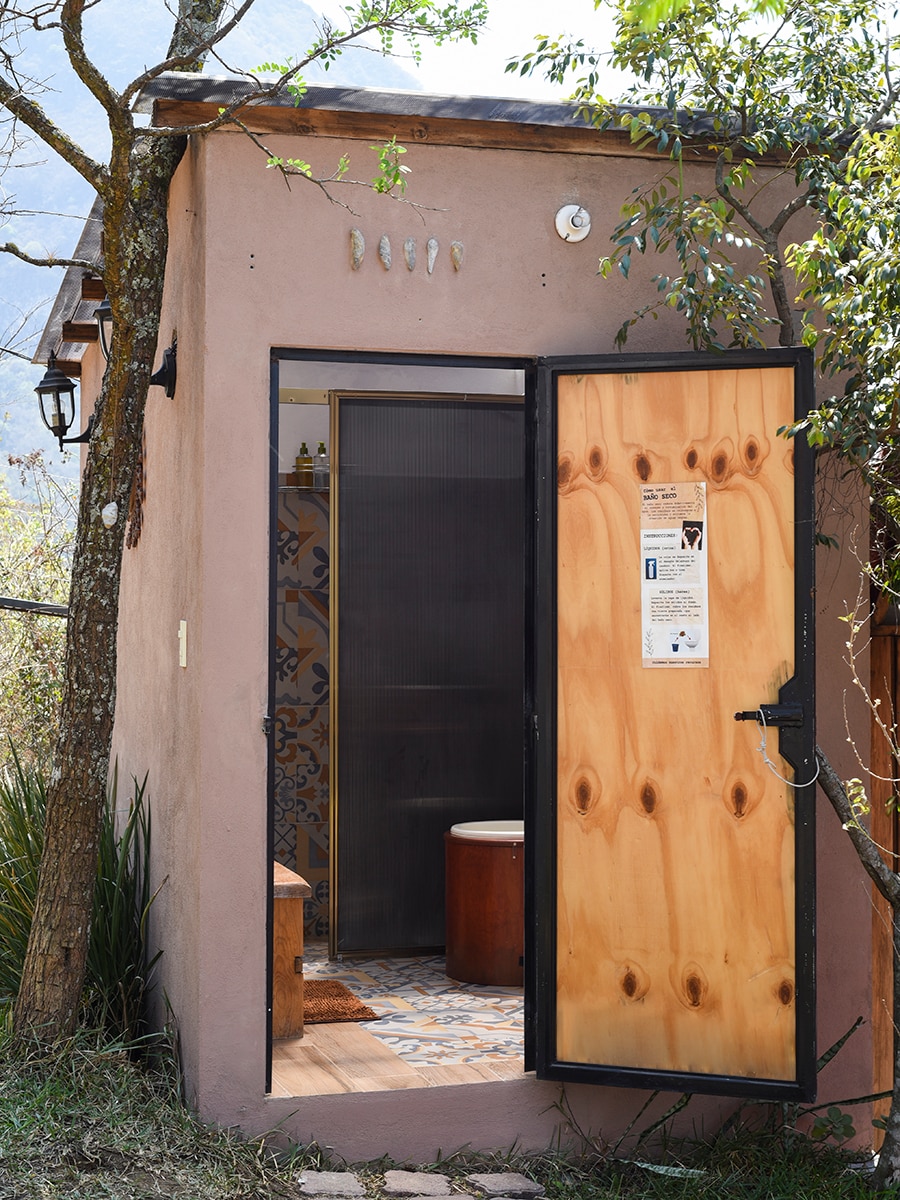 The entrance to a compact, rustic bathroom features a wooden door partially open, revealing tasteful tilework and a small round sink. Natural elements frame the doorway, while the surrounding area reflects a tranquil outdoor setting.