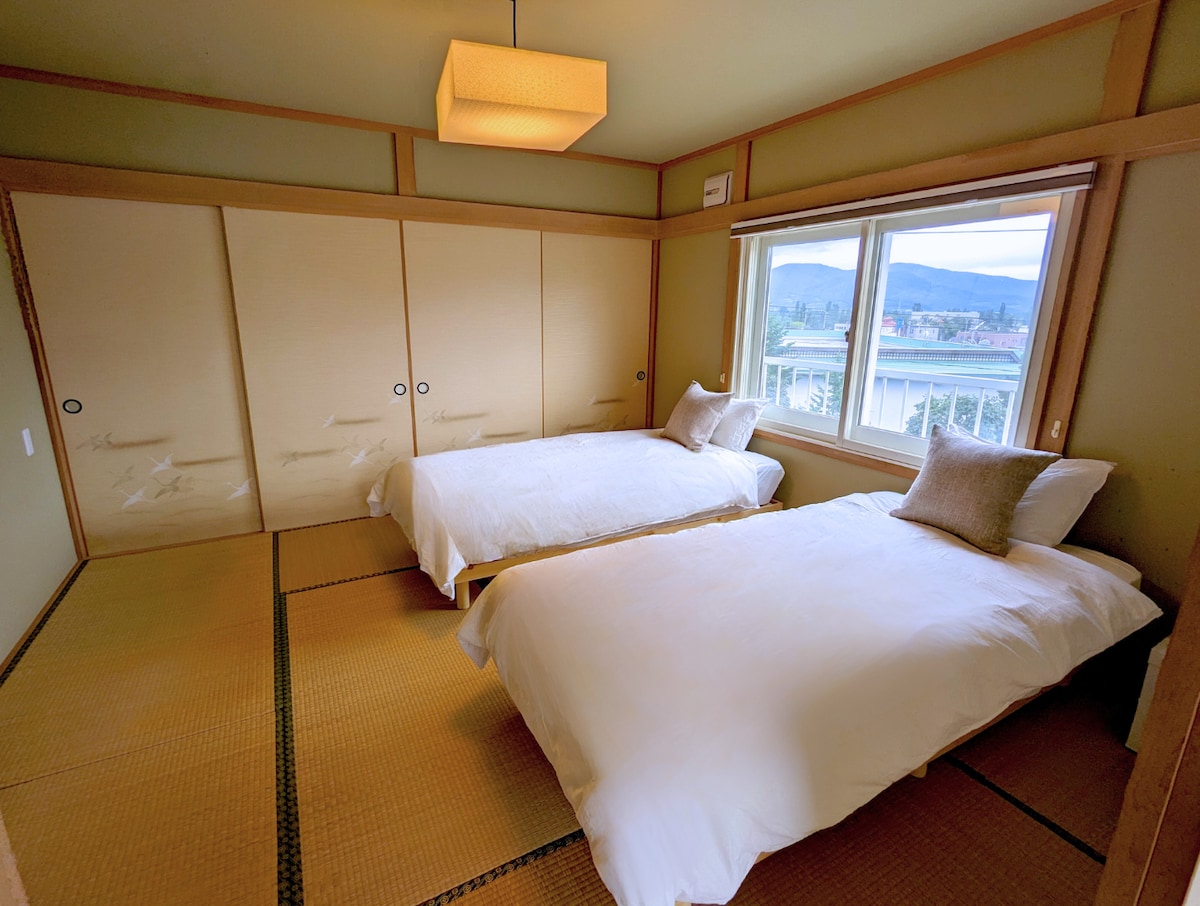 A serene bedroom features two spacious beds placed on tatami flooring, warmly lit by a central ceiling fixture. Sliding shoji doors allow natural light to enter, while a window offers a view of the surrounding landscape, creating a calm and inviting environment.