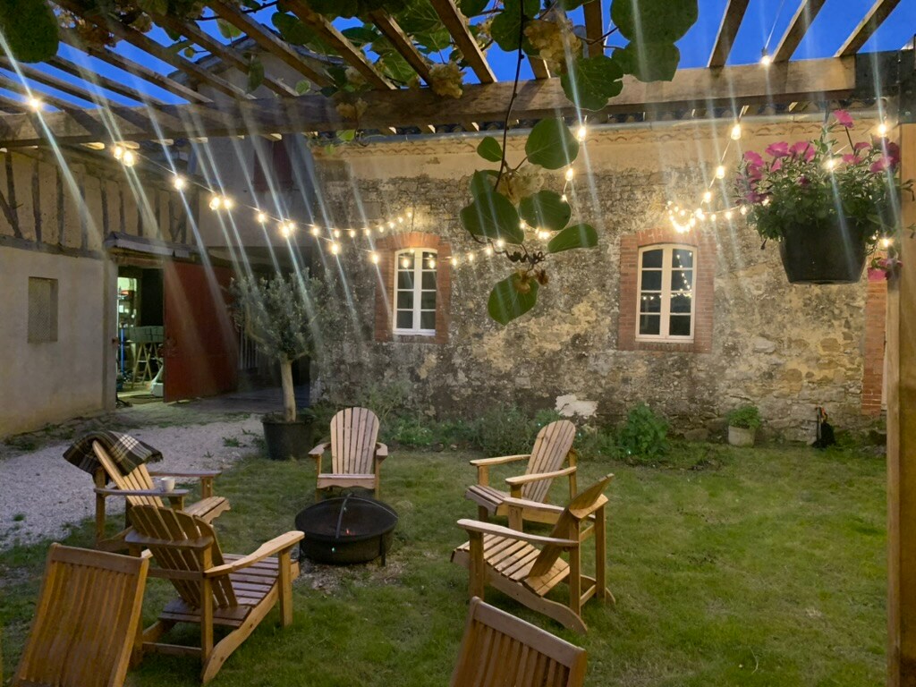 An enclosed courtyard space is framed by rustic wooden beams and illuminated by soft string lights. Several wooden adirondack chairs are arranged around a fire pit on a grassy area, with potted plants adding a touch of greenery against the historic stone walls.
