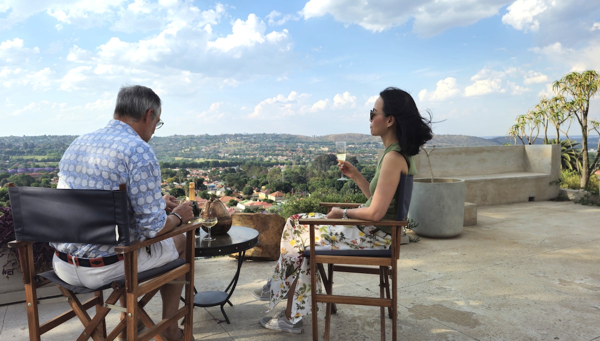 A serene outdoor seating area features two individuals enjoying refreshments. They are seated on wooden chairs around a small round table. The backdrop reveals a panoramic view of lush hills and a sprawling landscape under a bright sky, enhancing the relaxed atmosphere.
