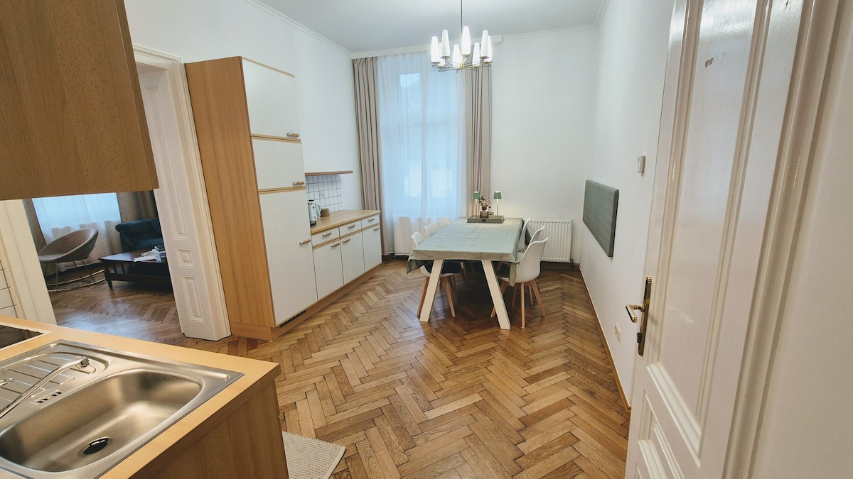 A bright dining area features a wooden table surrounded by four chairs, with a soft grey table runner on top. Natural light enters through a large window, illuminating the light wood cabinetry and herringbone patterned floor. An elegant light fixture hangs overhead.