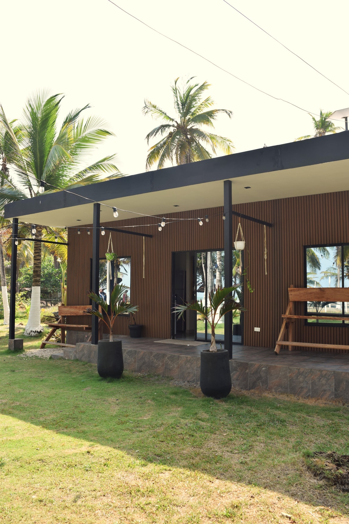 A stylish house is presented with a wooden exterior, featuring large glass doors that lead to a shaded porch. Potted plants are positioned at the entrance, alongside comfortable seating. Palm trees are visible in the background, complementing the inviting outdoor space.