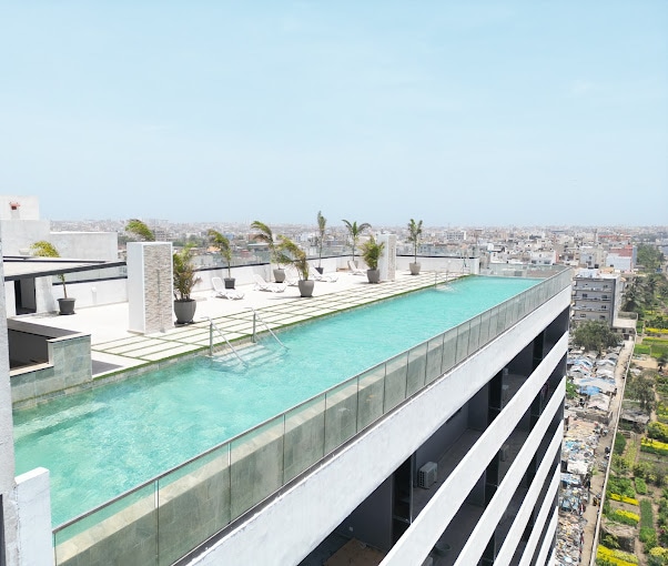 A rooftop terrace features a spacious infinity pool surrounded by potted palm trees. Loungers are positioned along the edge for relaxation, while a panoramic view of the city skyline extends in the distance under a clear blue sky.