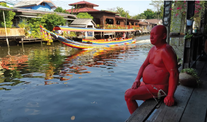 A person painted in red sits along a wooden platform beside a tranquil waterway. Colorful boats glide past, showcasing the vibrant atmosphere of the area. Lush greenery and local architecture line the banks, reflecting a lively connection to the surrounding Thai culture.