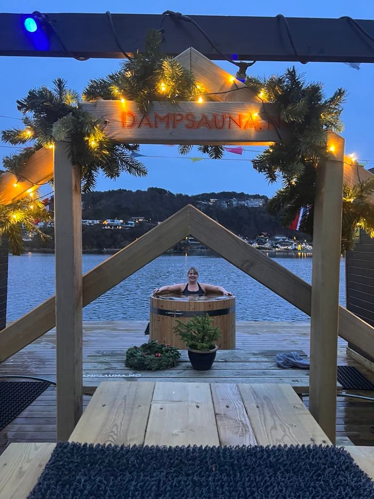 A wooden structure features a sign reading 'Hampsauna' adorned with festive lights and greenery. In the foreground, a small plant sits beside a hot tub, which is positioned on a dock overlooking the water, creating a serene atmosphere during twilight.