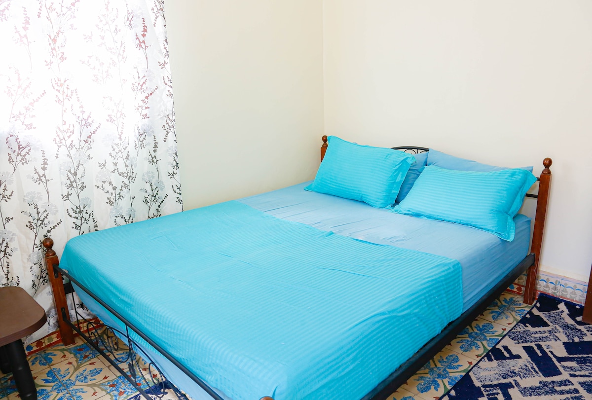 A comfortable sleeping area features a spacious bed adorned with textured blue bedding and matching pillows. A light curtain allows natural light to filter in, casting soft shadows on the walls. The patterned rug complements the decor, providing a warm touch to the space.