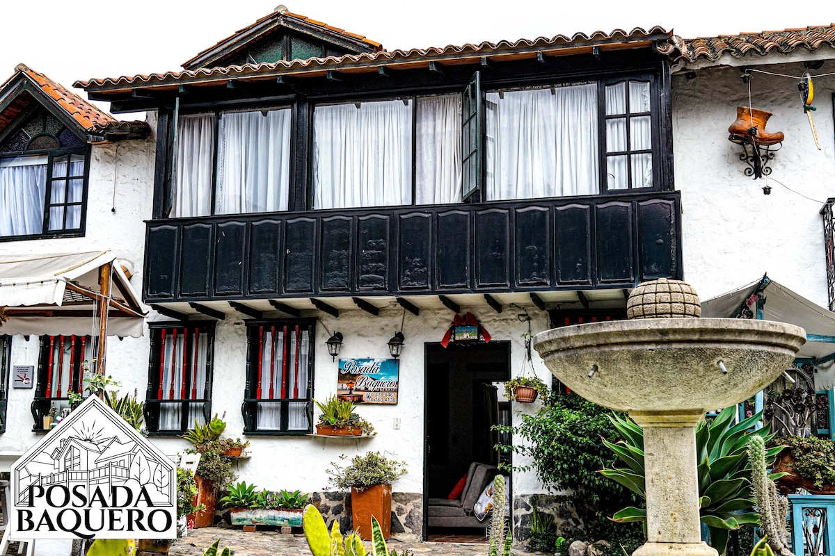 The two-story building features a charming white façade with black wooden accents. Large windows adorned with sheer curtains allow ample natural light. A decorative fountain sits in the foreground, surrounded by lush greenery and native plants, creating a tranquil outdoor setting.
