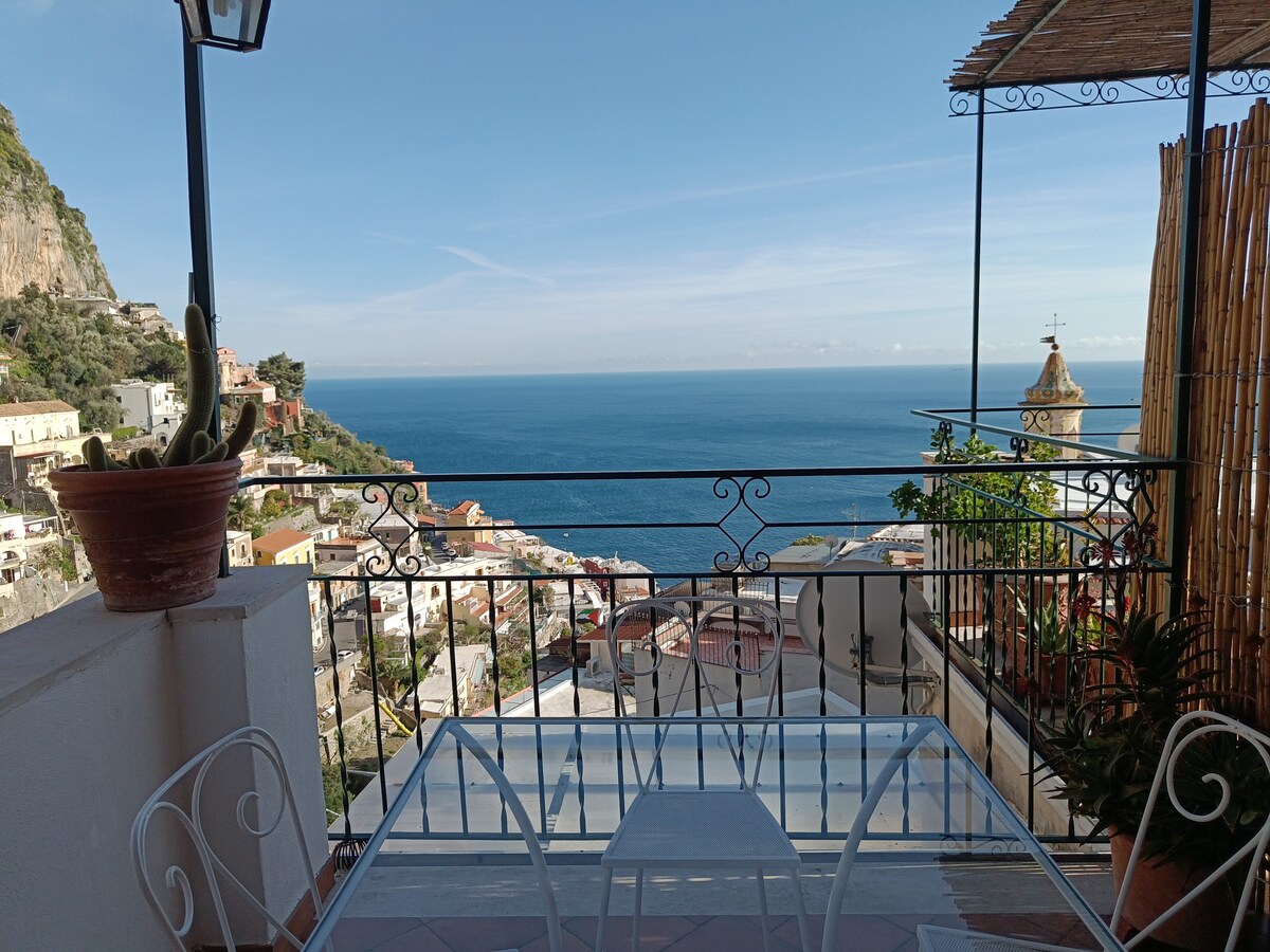 A spacious terrace is visible, featuring a glass table and white metal chairs. The expansive view showcases the blue waters of the bay and colorful homes along the hillside. A church bell tower can be seen in the distance, framed by lush greenery and a potted plant.