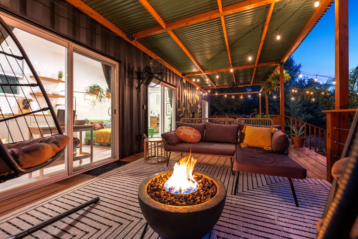 A spacious deck features cozy seating around a firepit, surrounded by string lights that enhance the outdoor ambiance. The interior of the container home is visible through large glass doors, providing a seamless connection between indoor and outdoor living spaces.