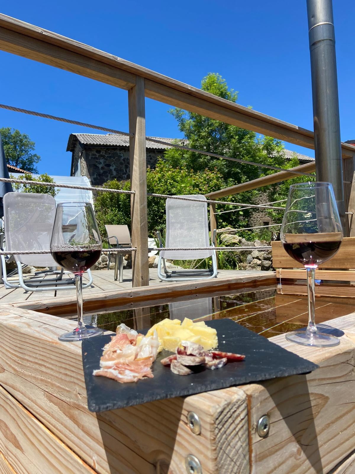 A wooden terrace showcases two glasses of red wine beside a slate platter with assorted meats and cheese. Sunlight illuminates the area, highlighting the surrounding greenery and comfortable lounge chairs positioned nearby.