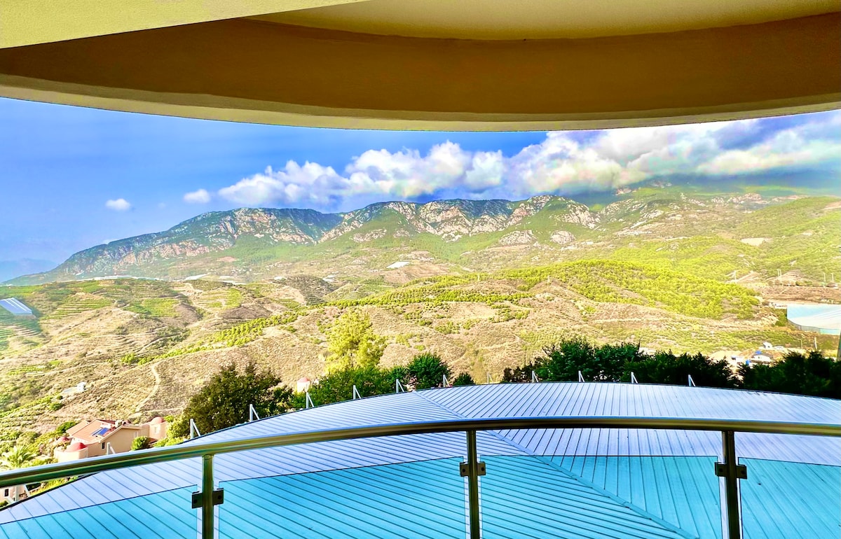 A panoramic view of lush green mountains is presented from a curved balcony. The landscape features rolling hills and a clear blue sky, enhancing the serene atmosphere. The railing of the balcony offers a sense of openness and connection to the natural surroundings.