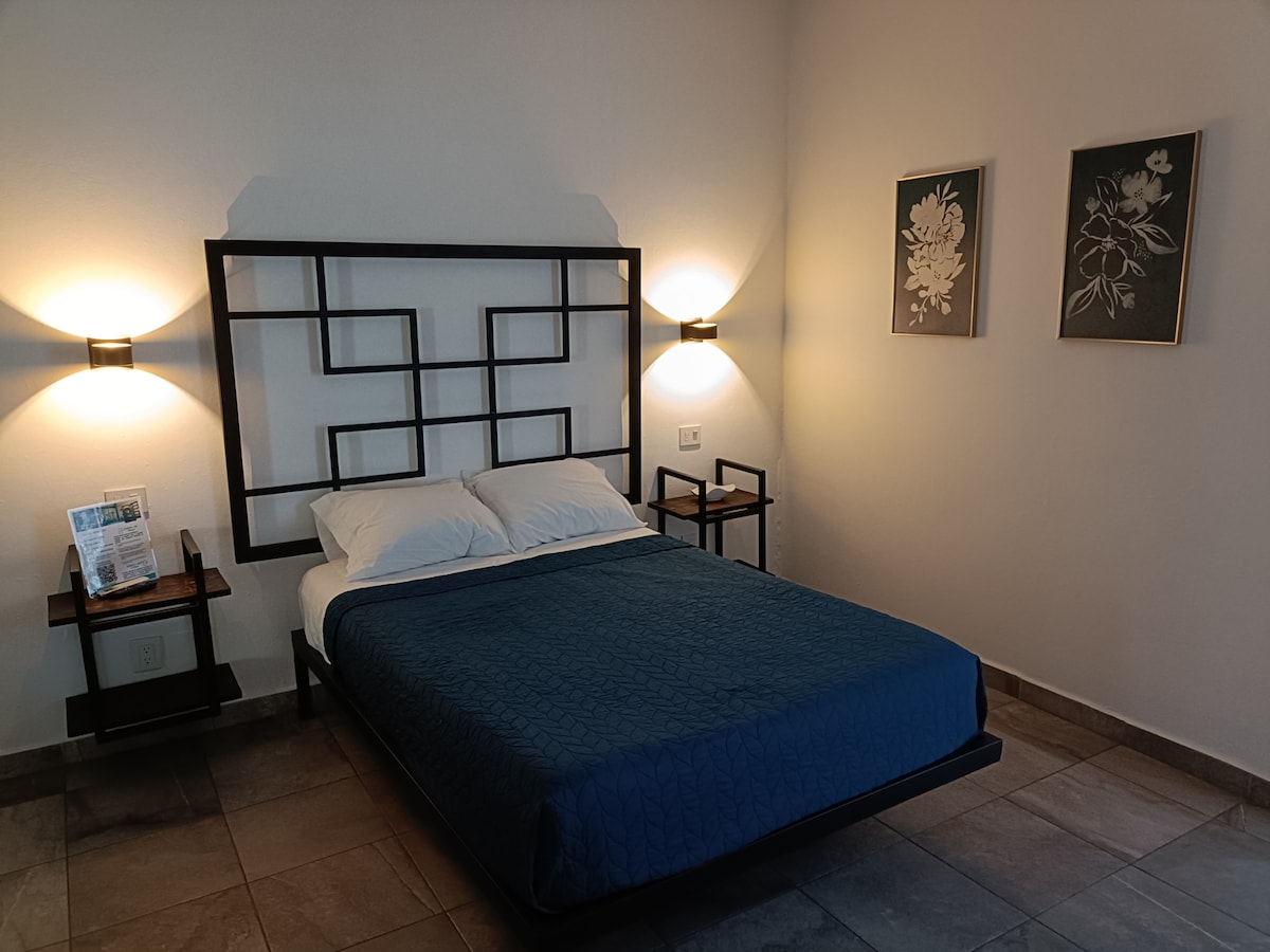 A spacious sleeping area features a modern bed with a dark blue coverlet. Flanking the bed are wooden nightstands with stylish lamps. Simple floral artwork is displayed on the wall, while soft lighting creates a serene environment.
