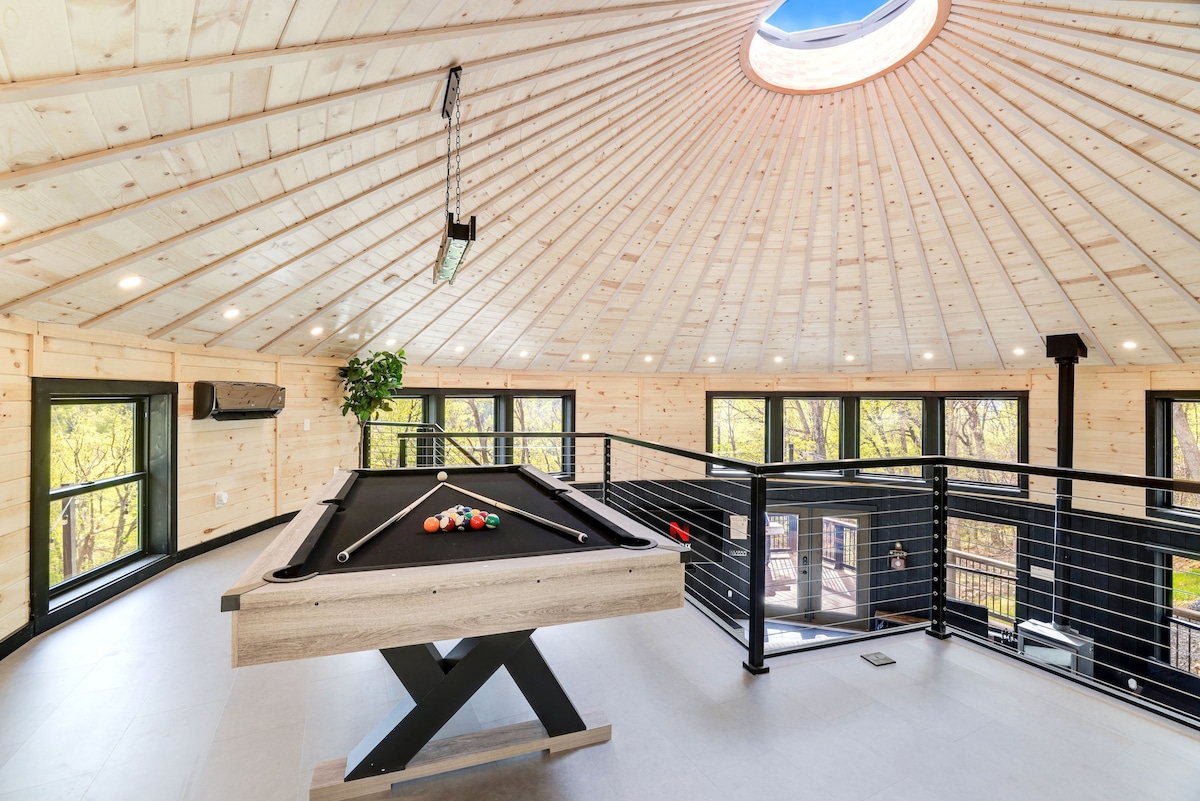The spacious loft area is highlighted by a circular skylight, allowing natural light to fill the room. A modern pool table occupies the center, surrounded by large windows that offer views of the surrounding forest. The wooden ceiling adds warmth to the contemporary design.