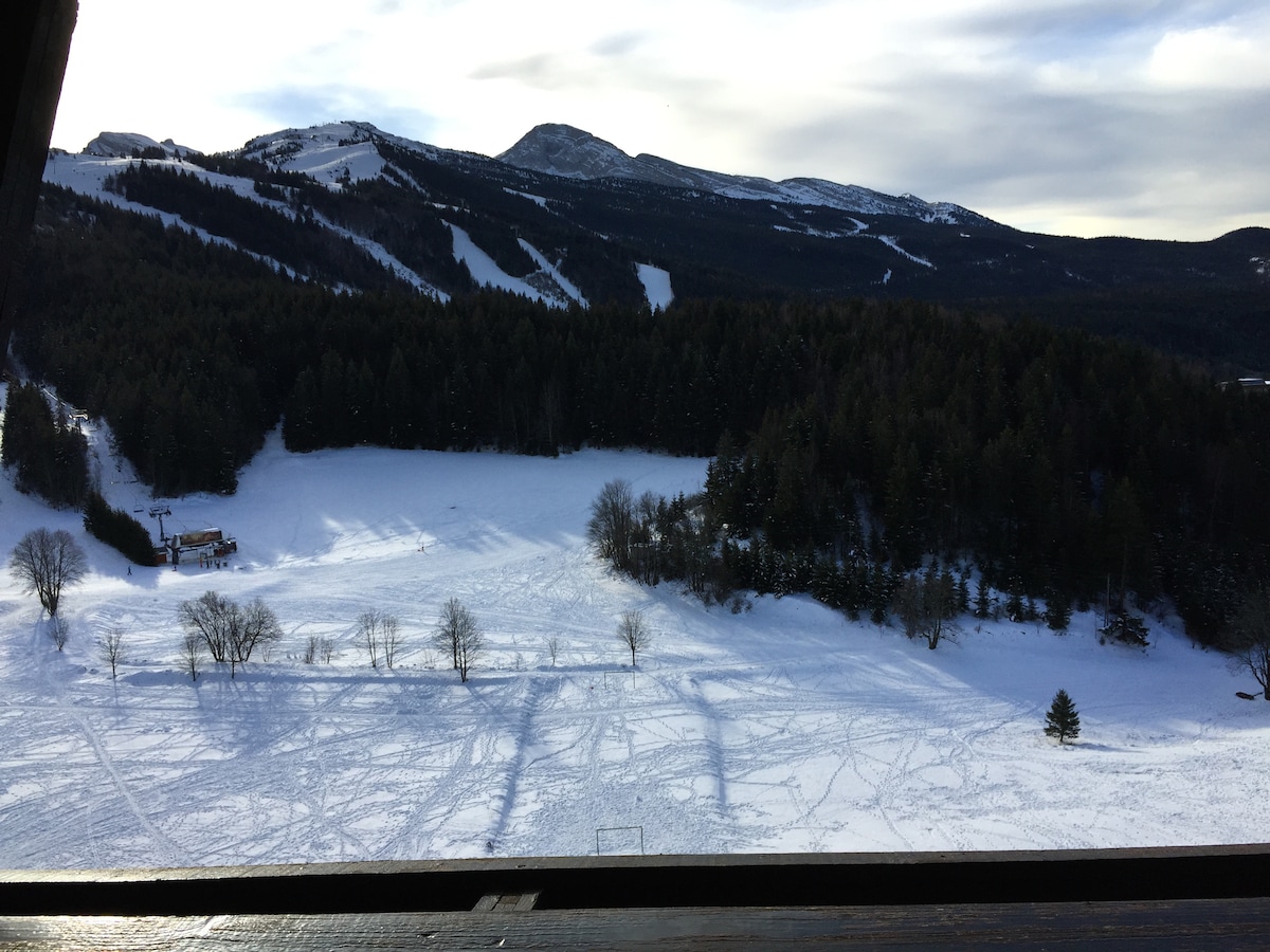 A snowy landscape unfolds, with gentle rolling hills and trees scattered across a white expanse. In the background, mountains rise against a cloudy sky, creating a serene winter scene. Tracks in the snow suggest recent activity, adding a sense of liveliness to the calm environment.