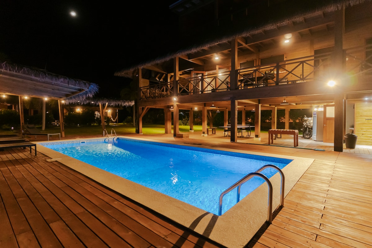 A well-lit swimming pool is set against a wooden deck, surrounded by natural green space. The pool features a gentle blue hue, with steps leading into the water. A shaded area with seating is visible, along with an upper balcony providing additional outdoor space.