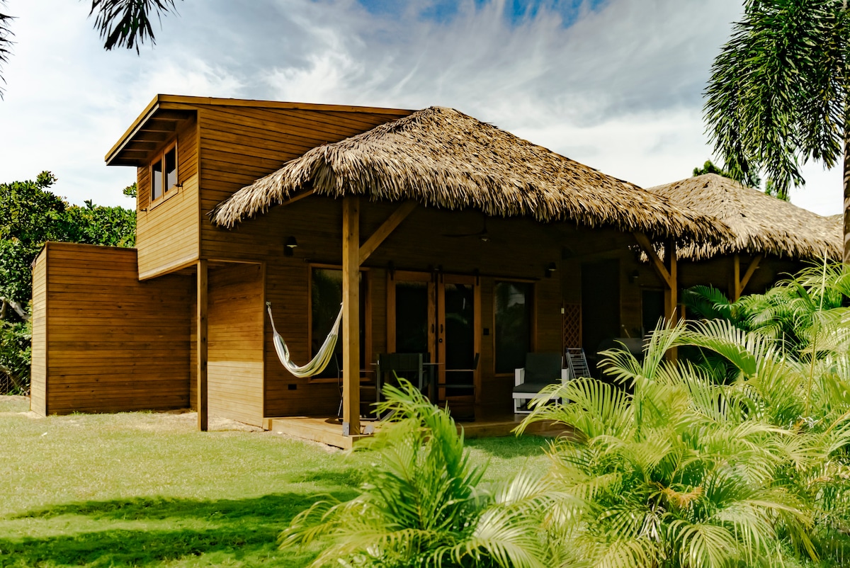 A wooden cabin is set amidst lush greenery, featuring a thatched roof and a spacious porch. A hammock is suspended between two posts, while comfortable seating arrangements are visible. The exterior showcases natural wood textures, blending harmoniously with the surrounding landscape.