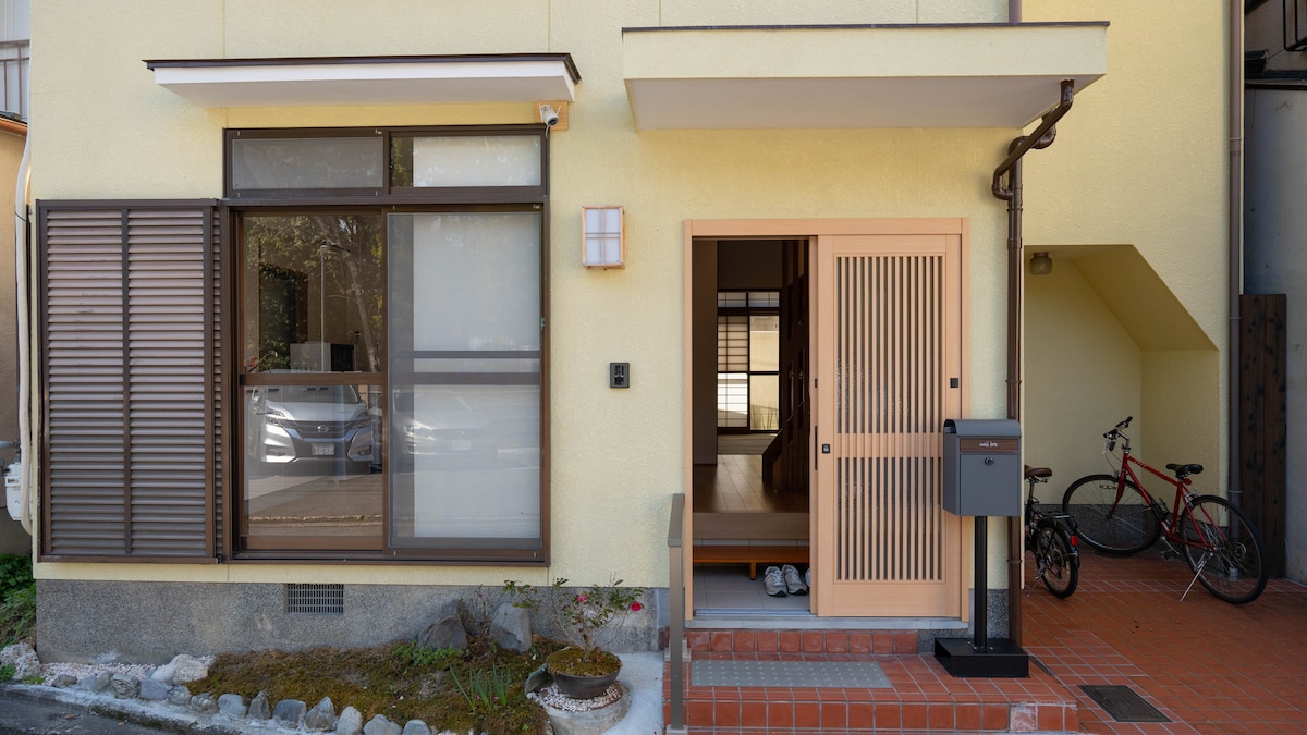 The exterior of a stylish residence is presented, featuring a yellow facade with large windows. A wooden door with a slatted design opens to reveal a glimpse of the interior. A bicycle is parked nearby, and a small garden adds greenery to the entrance area.