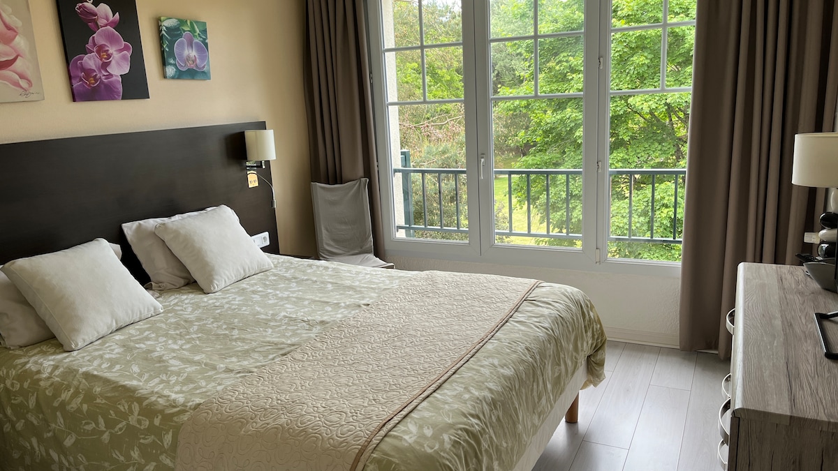 A spacious bedroom is furnished with a large bed, complemented by two bedside lamps. Large windows allow natural light to fill the room, offering a view of greenery outside. Soft floral artwork adorns the wall, enhancing the serene ambiance.
