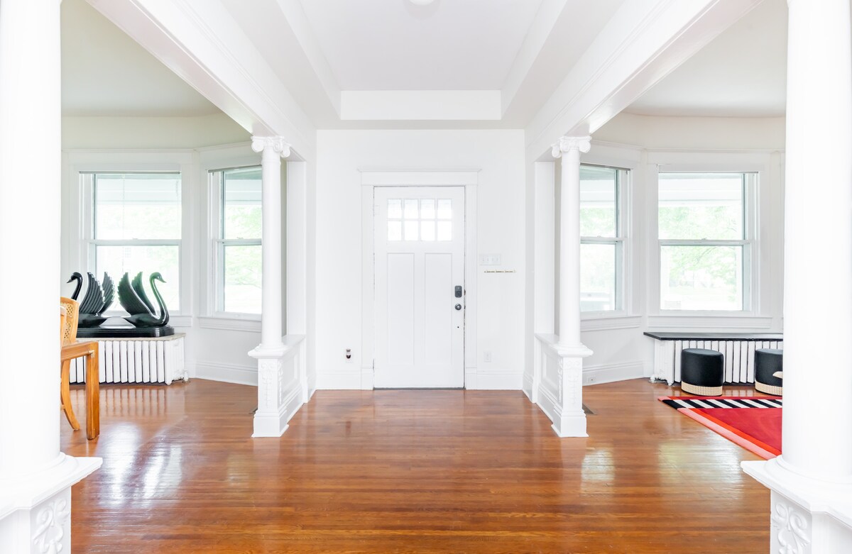 A bright and airy entryway features polished hardwood floors and tall, white columns. Light floods through large windows on either side, highlighting a mix of furniture. A decorative table with two black swan sculptures stands adjacent to a stylish black furniture piece in the background.