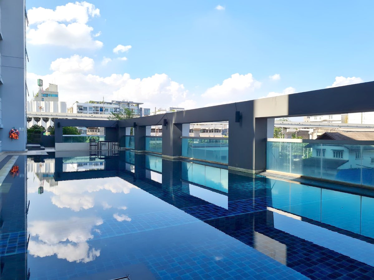 A tranquil swimming pool is situated on the rooftop, featuring a reflective surface with surrounding clear glass railings. The sky above is bright blue with fluffy clouds, providing a serene atmosphere. Sun loungers and a small seating area can be seen alongside the pool.