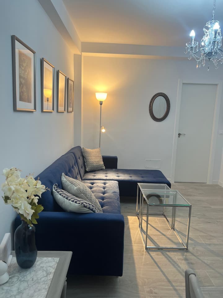 Salón-comedor con elegante sofá de respaldo ajustable y convertible en cómoda cama.  La hermosa lámpara de araña y los elementos decorativos en plateado añaden un brillo sutil y sofisticado, creando una atmósfera encantadora en este espacio.