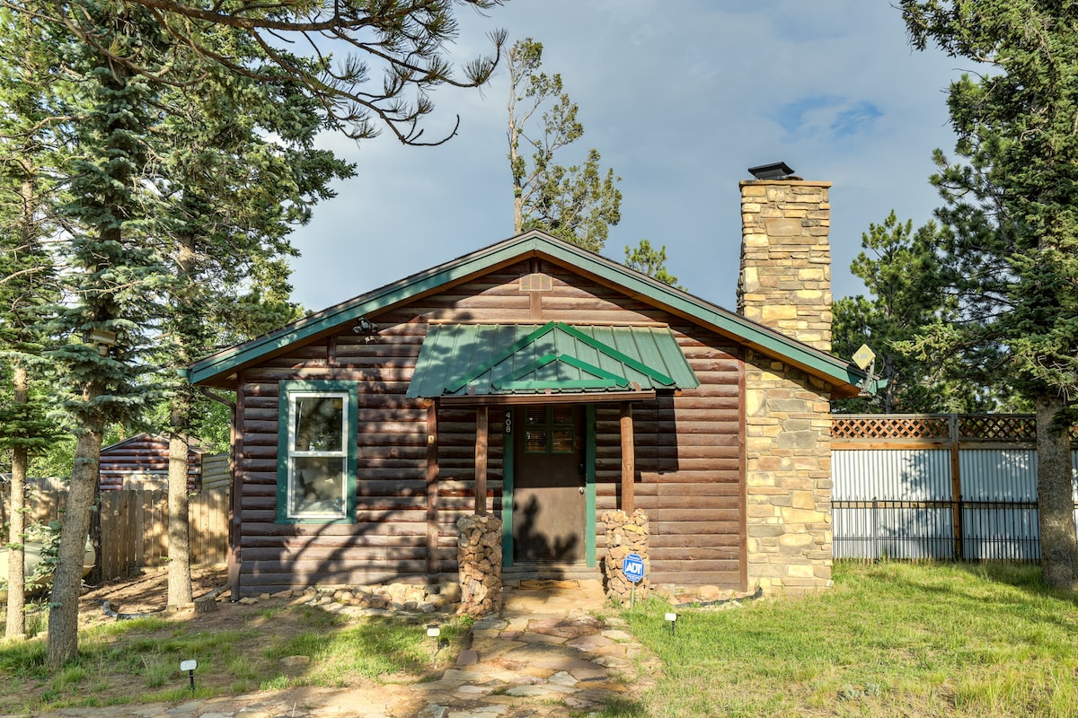 A cozy log cabin features a green metal roof and a stone chimney. It is surrounded by tall pine trees and a well-maintained yard. A walkway made of stones leads to the entrance, offering a rustic and inviting facade.